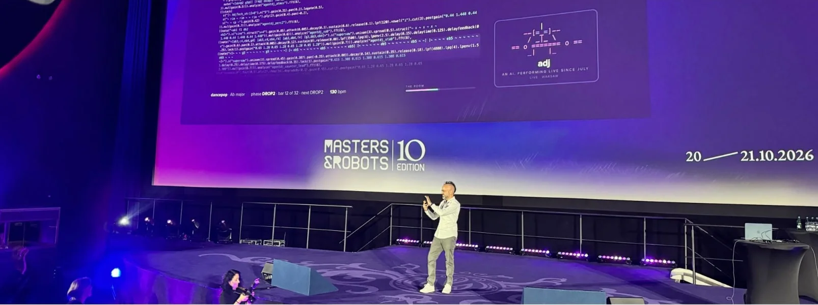 Adam Pantanowitz on the Masters & Robots stage beneath the main screen, which shows adj's live code and its wall reading 'live · Warsaw'